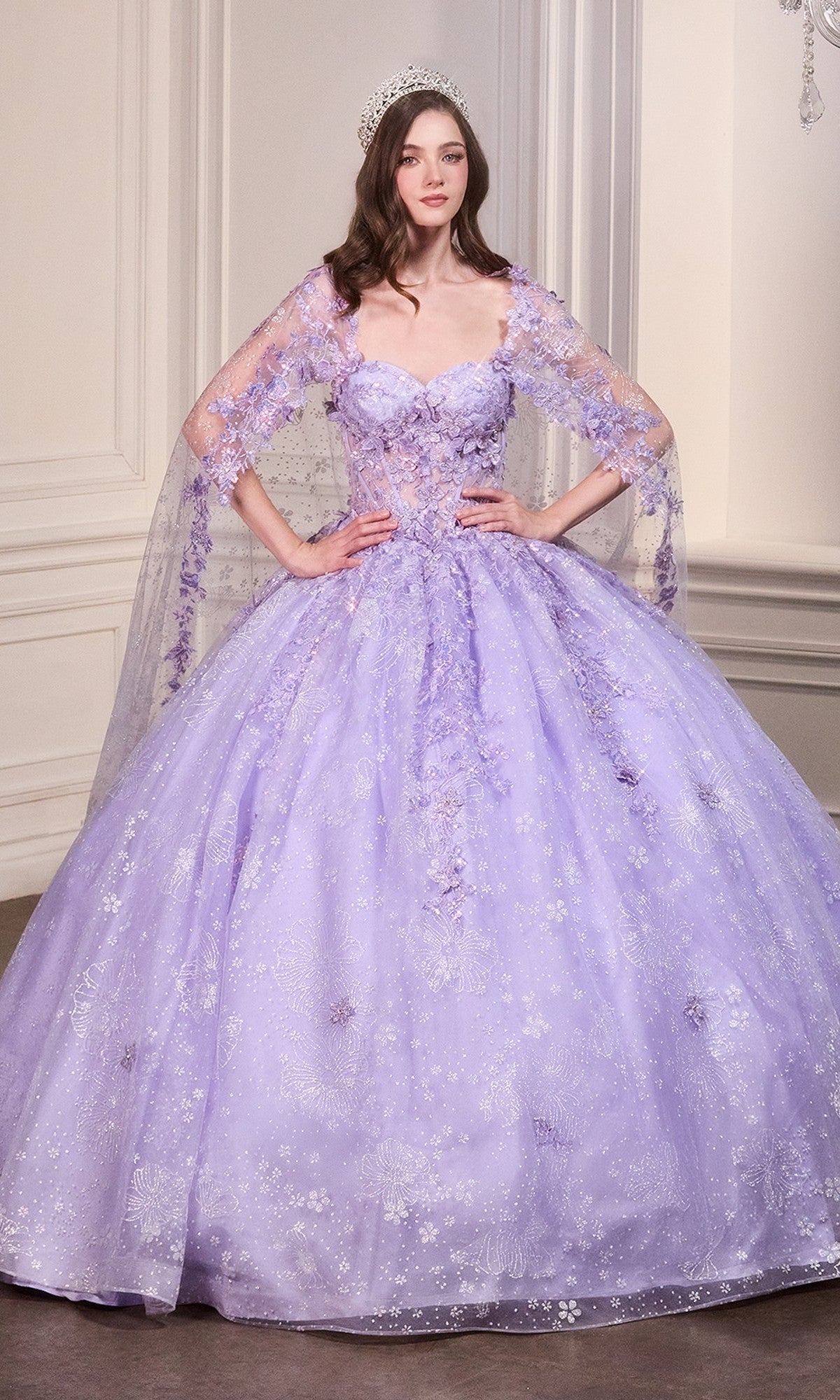 A woman wears the Ladivine Quinceanera Dress 15719, a light purple lace ball gown with floral accents and a sheer cape, styled with a jeweled headpiece, standing in an elegant room with white paneled walls.
